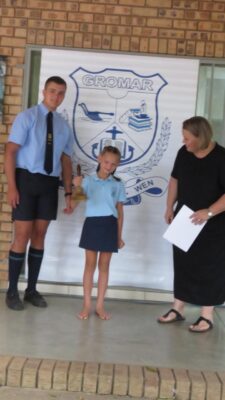 A young girl stands on stage holding a trophy, flanked by a smiling teenager and an adult holding papers. A school emblem is displayed behind them.