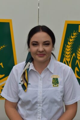 Smiling woman in a white shirt with a badge, wearing a striped scarf, stands in front of a board with green and yellow graphics, exuding professionalism.