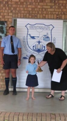 A young girl in a school uniform rings a brass bell, assisted by a woman. A man in a uniform stands nearby. They are in front of a banner reading "GROMAR."