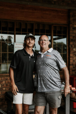 Two men stand side by side, smiling warmly. One wears a black polo and white shorts, the other a gray shirt and cargo shorts. They're in front of a brick building.