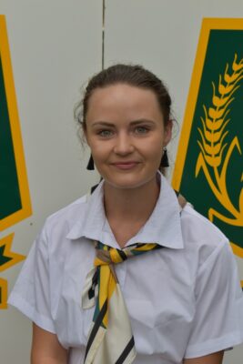 Woman in a white uniform with a patterned scarf poses against a backdrop featuring yellow and green agricultural logos. She has a calm expression.