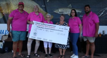 Six people stand smiling with a large ceremonial check for 150,000 rand, wearing matching pink shirts, in front of a fishing-themed banner.