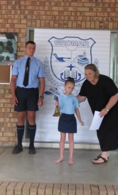 A young girl in a blue uniform rings a bell, guided by a smiling woman holding a paper. A boy in uniform stands next to a school emblem backdrop.