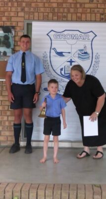 A young boy proudly holds a bell between a smiling woman and a taller student in uniform in front of a school emblem on a brick wall, conveying a celebratory tone.