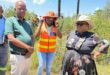 Four people stand outdoors by a concrete structure, next to tall grass. One wears an orange safety vest and hat. The mood is casual and conversational.