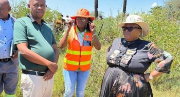 Four people stand outdoors by a concrete structure, next to tall grass. One wears an orange safety vest and hat. The mood is casual and conversational.
