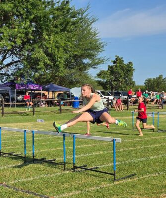 Young athlete mid-air over a hurdle in a race; she wears a light green tank top and navy shorts. Spectators and trees are in the background, with a sunny, energetic atmosphere.