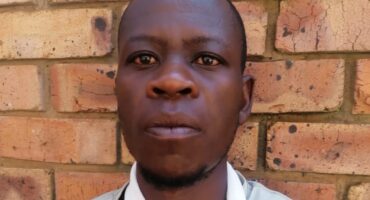 A person with a serious expression stands in front of a rustic brick wall. The image conveys a tone of contemplation or determination.