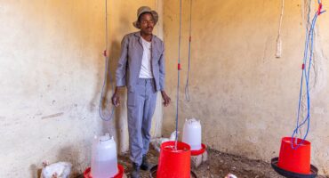 A man in a gray suit and hat stands in a poultry shed with white chickens. Red and white water dispensers hang from the ceiling, creating a hardworking, rural atmosphere.