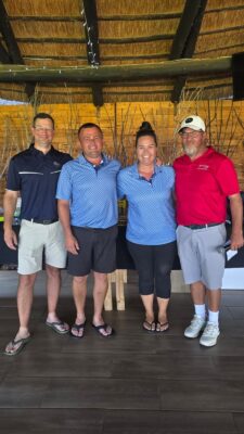 Four people stand arm in arm under a thatched canopy, smiling warmly. They are casually dressed, with two wearing matching blue shirts and another in a red shirt.