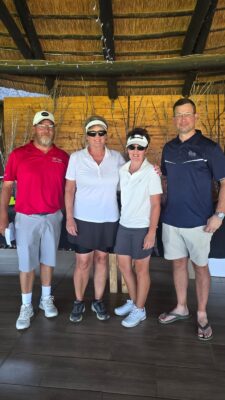 A group of four people stand under a thatched roof. Two women in golf attire are flanked by two men, one in a red polo, the other in navy. Smiling, casual atmosphere.