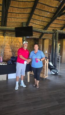 A man and woman smiling and shaking hands in a rustic venue with a wooden ceiling. The man wears a red shirt and cap; the woman holds a pink item.