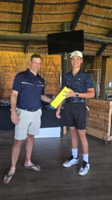 Two men are standing indoors with wooden walls and a thatched roof. One man hands a bright yellow rectangular item to the other. Both are smiling, creating a friendly atmosphere.