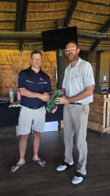 Two men stand in a wooden building, smiling and shaking hands. One holds a green trophy. They are dressed in casual golf attire. The mood is celebratory.