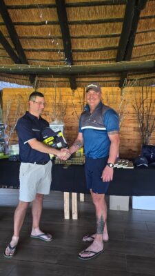 Two men smiling and shaking hands indoors. One holds a boxed gift. Background includes a rustic wooden wall and dark-draped table with items.