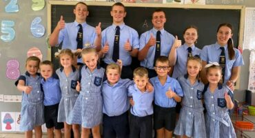 A group photo in a classroom shows children and older students in uniform, smiling and giving thumbs up. The atmosphere is cheerful and positive.