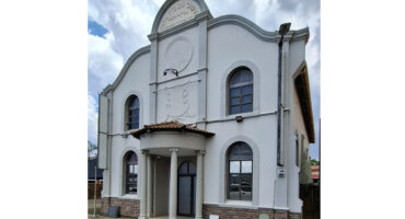 Historic synagogue with a white facade, arched windows, and a decorative entryway with columns. The sky is partly cloudy, conveying a serene atmosphere.