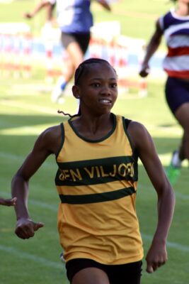 A young athlete in a yellow and green jersey runs determinedly during a track race. The background shows blurred competitors and a grassy field.