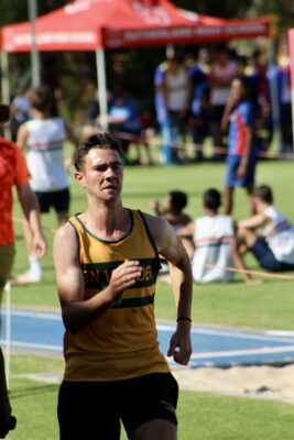 A male athlete in a yellow and black tank top is sprinting outdoors on a sunny day. A red tent and other runners are seen in the blurred background.