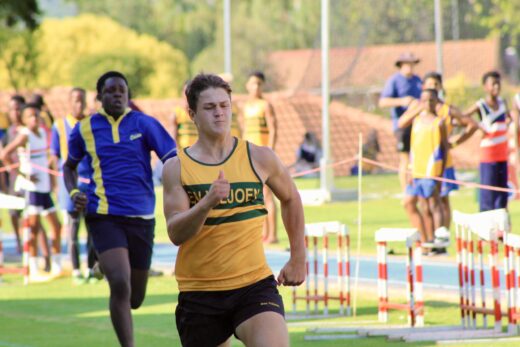 Male athlete in yellow tank top sprints confidently on a track, with competitors and hurdles in the background on a sunny day. Focused and determined.