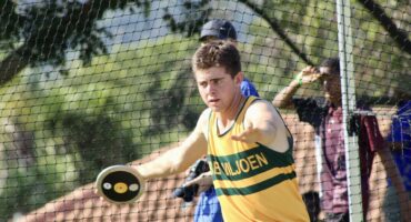 A male athlete in a yellow and green jersey throws a discus in a netted field. His expression shows focus. Background includes trees and observers.
