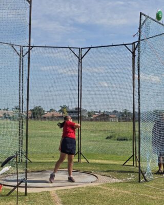A person in a red shirt and black shorts throws a discus in an outdoor sports field, surrounded by safety nets, under a clear blue sky.