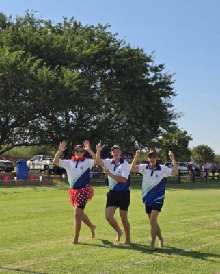 Three people in matching shirts jog joyfully on a sunny field. One wears a red tutu and a hat. Trees and cars are in the background.