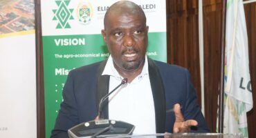 A man stands at a podium, speaking. He wears a dark suit and white shirt. Behind him is a banner with visions and missions text and a logo.