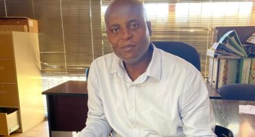 A man in a white shirt sits at a desk in an office with a calm expression. Behind him are files, shelves, and blinds filtering sunlight.