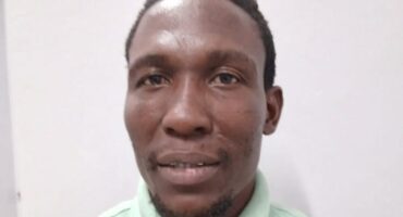 A man in a light green shirt against a plain white background, looking directly at the camera with a neutral expression. The lighting is even and soft.