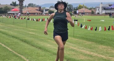 A person is running barefoot on a grassy field, wearing a black hat and sleeveless top. Flags line the track. The scene is bright and energetic.