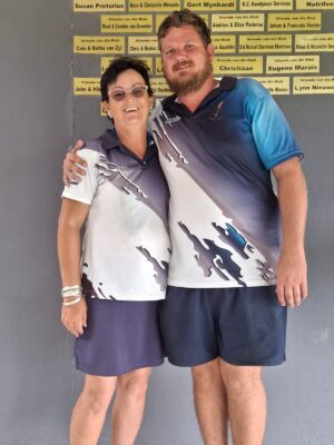 A smiling woman and man stand arm-in-arm, wearing matching sports shirts with abstract designs. Yellow name plaques are visible in the background.