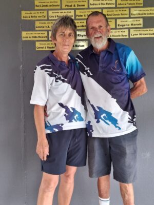 An elderly couple stands close together smiling warmly. They wear matching sports shirts with blue and white patterns against a gray wall featuring name plaques.