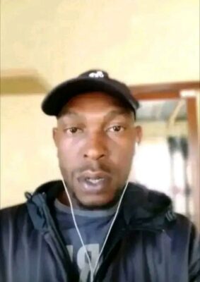 A person wearing a black cap and jacket is indoors, staring directly at the camera. They have earbuds in and a neutral expression.