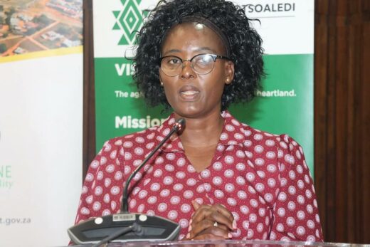 A woman in glasses and a red patterned blouse speaks at a podium, appearing focused and professional. Two banners with green accents are in the background.