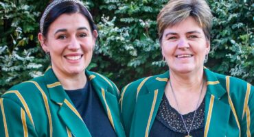 Two smiling individuals wearing green blazers with yellow stripes stand side by side against a backdrop of lush greenery, conveying a cheerful and positive mood.
