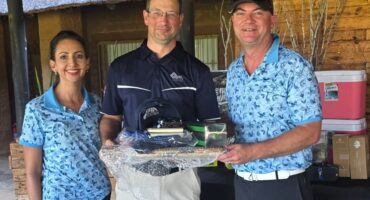 Three people smiling, two in light blue shirts with patterns, holding a prize basket wrapped in plastic. They're outside near a wooden building, conveying a cheerful mood.