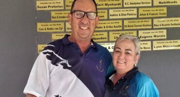 A smiling man and woman in sports shirts stand together in front of a wall with donor plaques. The mood is warm and friendly.