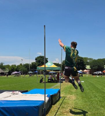 A male athlete in a green uniform is mid-jump over a high jump bar on a sunny day. The scene is set at an outdoor sports field with trees, tents, and blue skies.