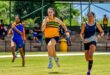 Three female athletes sprint on a grassy track, captured mid-stride. They exude determination, with spectators visible in the shaded bleachers behind them.