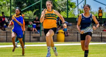 Three female athletes sprint on a grassy track, captured mid-stride. They exude determination, with spectators visible in the shaded bleachers behind them.