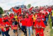 A large group of people wearing red and holding signs march in a street protest. The atmosphere is energetic and focused, with various slogans visible.