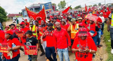 A large group of people wearing red and holding signs march in a street protest. The atmosphere is energetic and focused, with various slogans visible.
