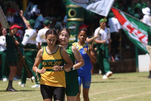 A group of female athletes in school uniforms run on a field. Spectators with colorful flags cheer in the background. The scene is lively and competitive.