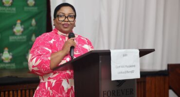 A person in a pink floral dress speaks at a podium with a microphone. Behind them is a green banner with indistinct text and logos. The atmosphere is formal.