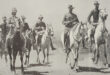 A group of six soldiers on horseback ride in formation on a dusty trail. They wear early 20th-century military uniforms with hats, exuding determination.