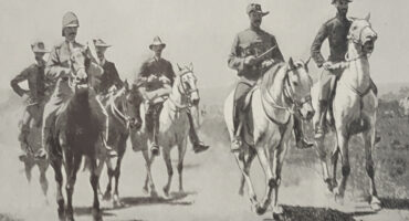 A group of six soldiers on horseback ride in formation on a dusty trail. They wear early 20th-century military uniforms with hats, exuding determination.