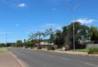 Wide street lined with palm trees under a clear blue sky. Sidewalks and light poles are visible; houses in the background create a tranquil suburban scene.