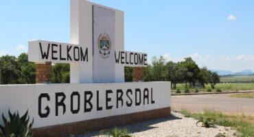 A white welcome sign reads "Welkom / Welcome to Groblersdal," set against a rural landscape with trees and distant hills under a clear blue sky.