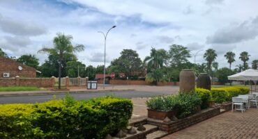 Paved patio with white chairs and tables, lush green shrubs in planters, palm trees in the distance under a cloudy blue sky. Calm suburban street.